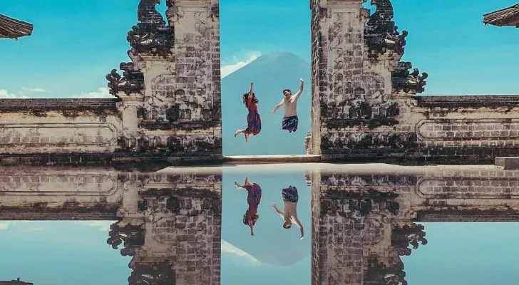 Two people jumping between two carved stone pillars with the scene mirrored in a reflection below
